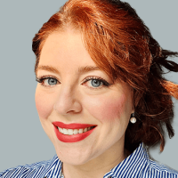 Close-up portrait of a smiling woman with red hair, blue eyes, wearing a striped shirt and pearl earrings.