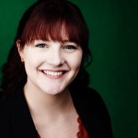 Portrait of a smiling woman with dark red hair and bangs, wearing a black blazer, against a green background.