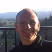 Smiling man wearing a dark jacket standing on a balcony with hills and mountains in the background.