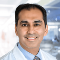 Smiling male doctor in a white coat and blue shirt in a bright clinical setting.
